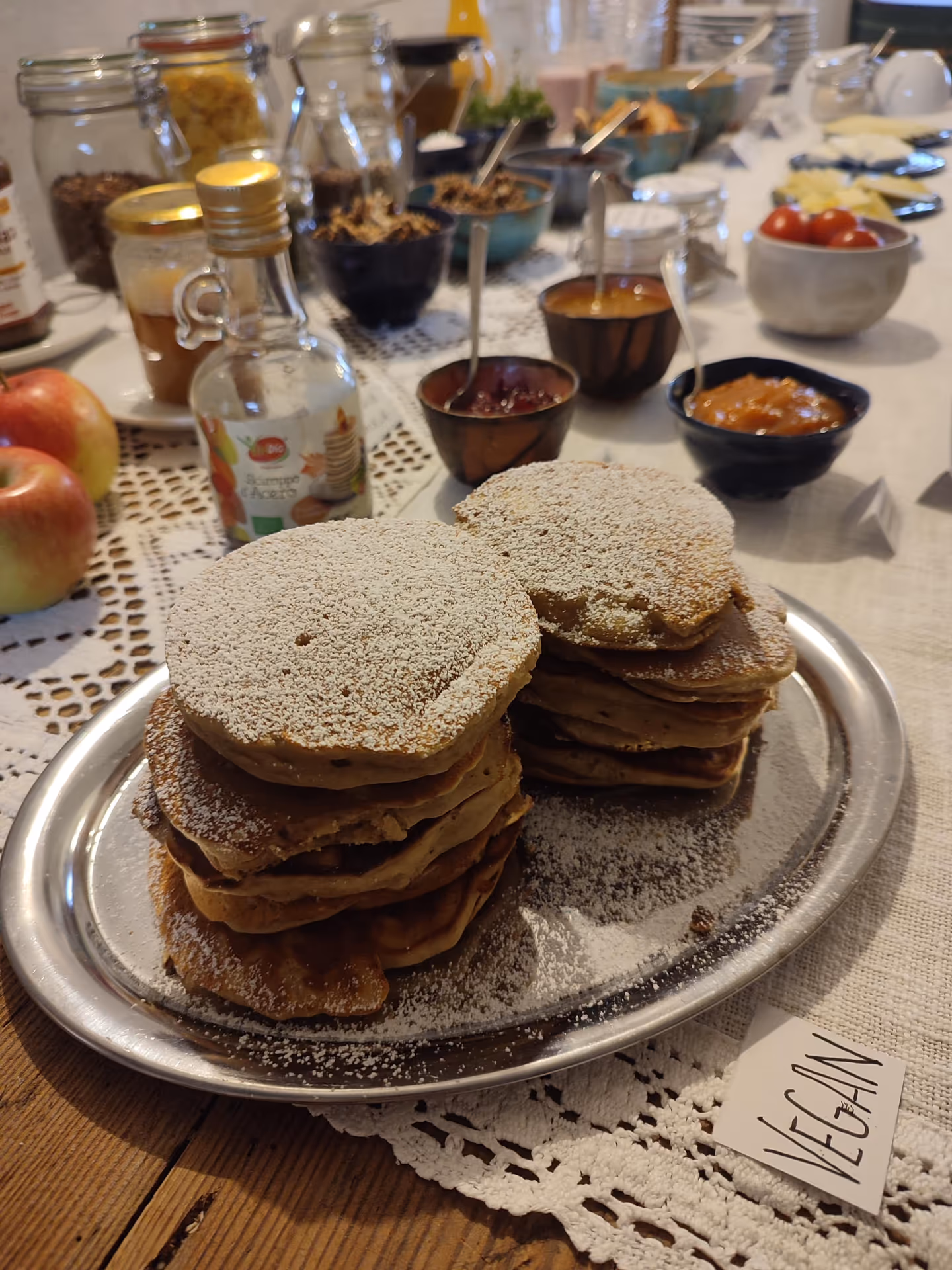 A plate of vegan pancakes with icing sugar, beside spreads, muesli and fruit